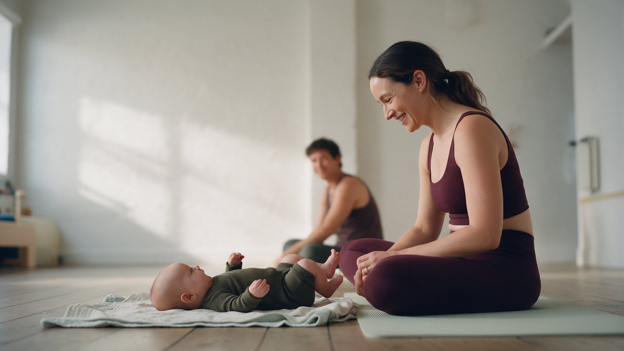 Illustrative image of a mother seated on a Pilates mat, smiling at her baby on a separate blanket